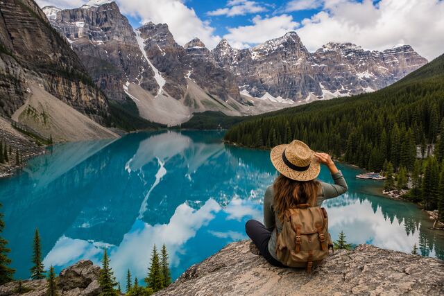 Turquoise Lake Louise surrounded by pine forests and snow-capped Canadian Rockies with a hiker overlooking the mountain landscape.