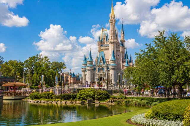 Cinderella Castle at a sunny Orlando theme park with families, greenery, and a reflective lagoon.