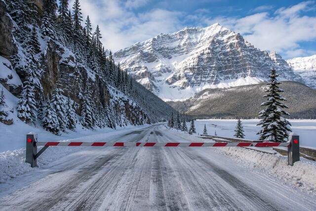 Snow-covered Banff winter road blocked by a barrier with mountains and a frozen lake in the background
